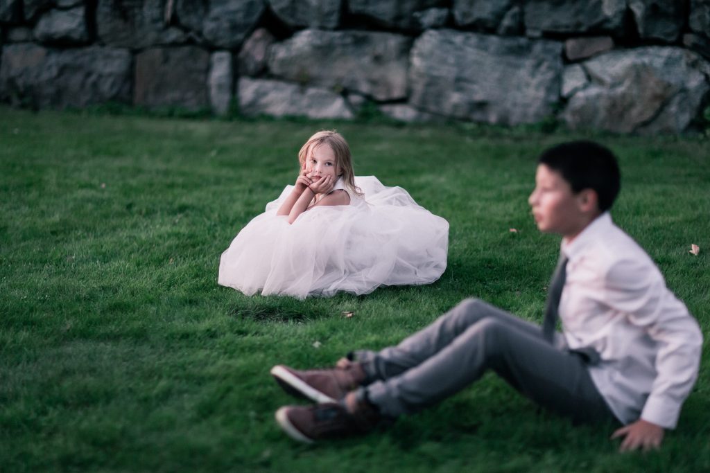 flower girl sitting in grass looking at a boy in a tie 