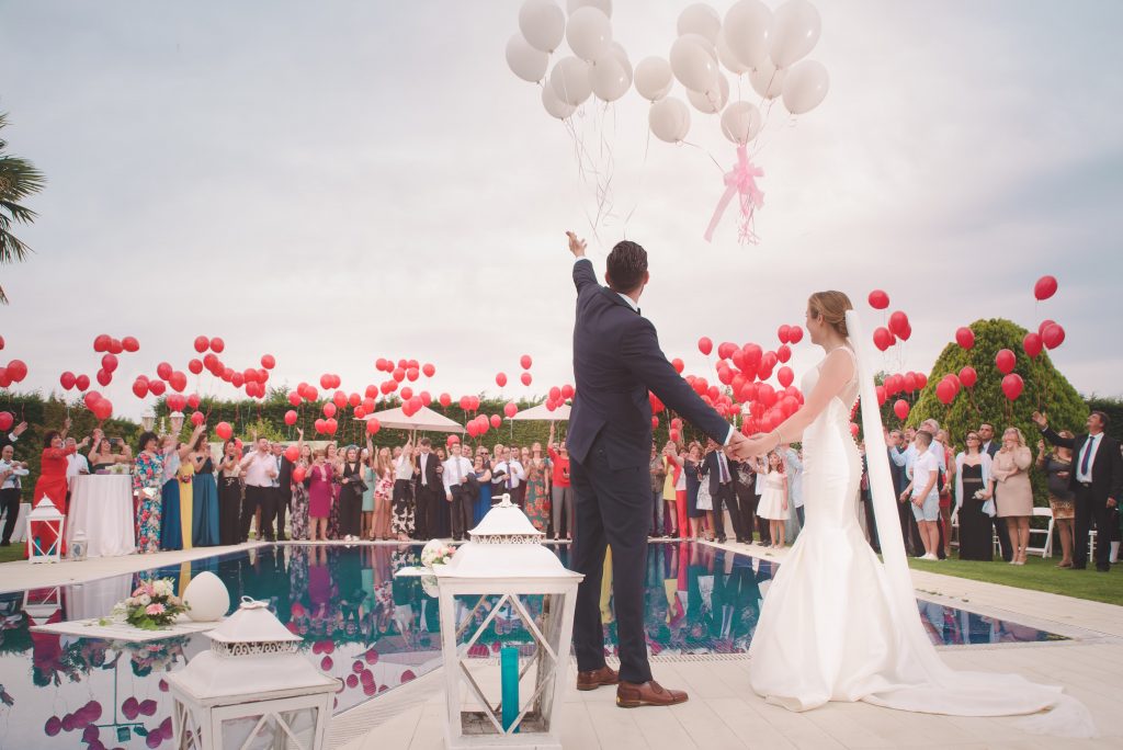 bride and groom next to a pool with their guests holding red balloons