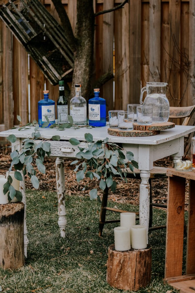 rustic bar set up with a wooden desk as the bar, liquor bottles, water pitcher, decorative wood slabs, candles, and eucalyptus garland